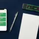 A flat-lay overhead shot of a dark navy workbench with a smartphone displaying a job scheduling app, a mechanical pencil, a closed notepad, and a spirit level, representing job management tools for UK tradespeople.