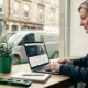 Does a small business need a website? This female tradesperson in a navy work jacket thinks so, reviewing her business website on a laptop with a UK high street visible behind her.